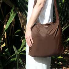 A person holding a Burnt Umber SYZY Tetra Tote Bag with multiple straps, surrounded by plants, against a natural background.